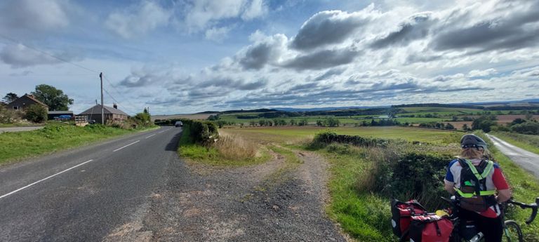 Looking east north east along the B6360 from the junction, with another dark structure, Smailholm Tower, visible on a hill top..