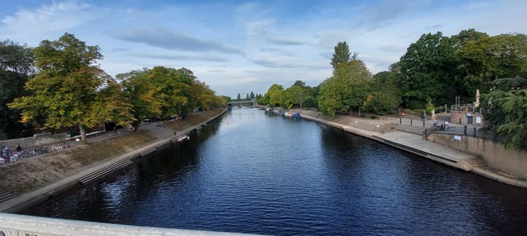 View up the River Ouse from the 1863 Lendal Bridge, crossing it north east to south west. York railway station is left of the tress on the left. The National Railway Museum is to the right of those trees on the left bank.