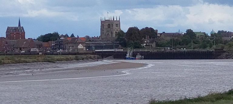 King's Lynn on the east side of the River Great Ouse - seen from the cycleway in.   From left: Spire of unseen 13th C St Nicholas Chapel, England's largest "chapel of ease",  hidden the Granary in Bakers Lane, built in 1900 to make linseed cake for cattle feed, now flats, and to the right St Margaret's church King's Lynn aka King's Lynn Minster., built 12th-15th century  St Nicholas's chapel: https://commons.wikimedia.org/w/index.php?search=st+nicholas+chapel+king%27s+lynn&title=Special:MediaSearch&go=Go&type=image         Granary: https://commons.wikimedia.org/wiki/File:Granary_King%27s_Lynn.JPG       Minster: https://en.wikipedia.org/wiki/King's_Lynn_Minster