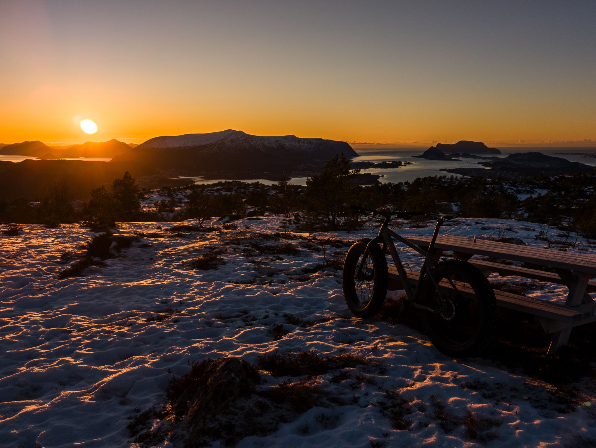 Fatbike ved en rastebenk i snødekt terreng med solnedgang over fjell og hav