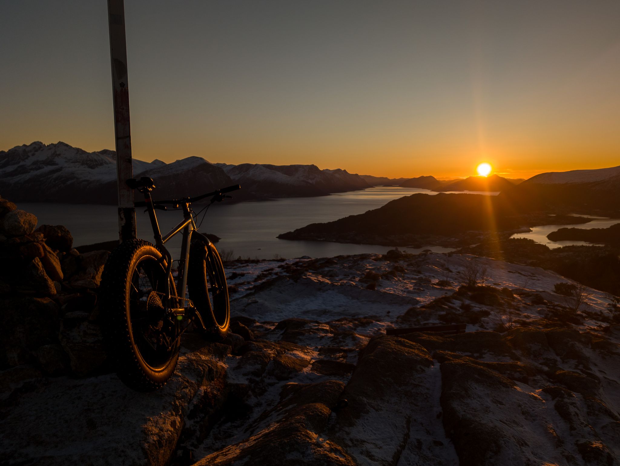 Fatbike ved en høy markør i snødekt toppterreng med fjord og fjell i solnedgang